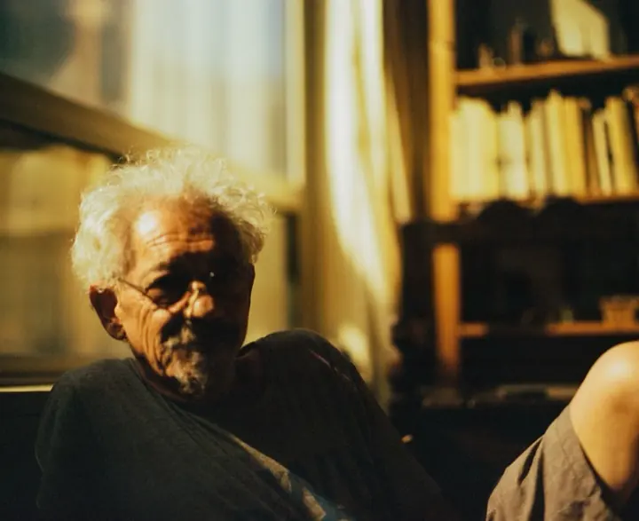 An older person with wild white hair, glasses and a grey beard sits in warm yellow light in front of bookshelves and curtains, wearing a dark top.
