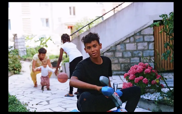 A teenager in a black t-shirt sits on a blue scooter in a paved garden with pink hydrangeas, a toddler with a basketball and two adults behind.