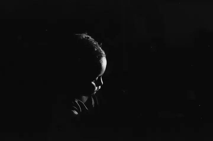 Very dark black and white profile of a child's head looking down, lit faintly from one side against a black background.