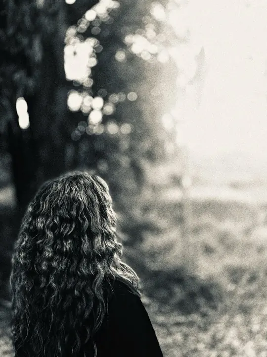 Black and white photo of a person with long curly hair seen from behind, facing a blurred sunlit tree and field.