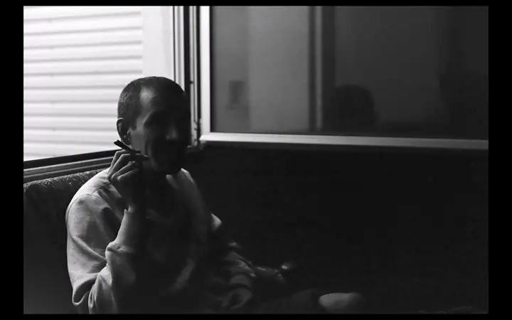 Black and white photo of a person in a light sweatshirt sitting by a window, holding a cigarette near their mouth in a dim room.