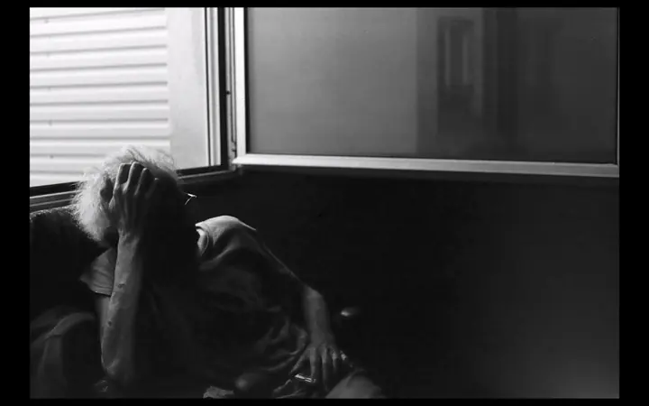 Black and white photo of a person with white hair holding their head in their hands, seated beside a window in a dim room.