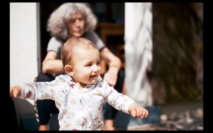 A baby in a floral top reaches out with arms wide, smiling, while an older person with curly grey hair sits behind in sunlight.