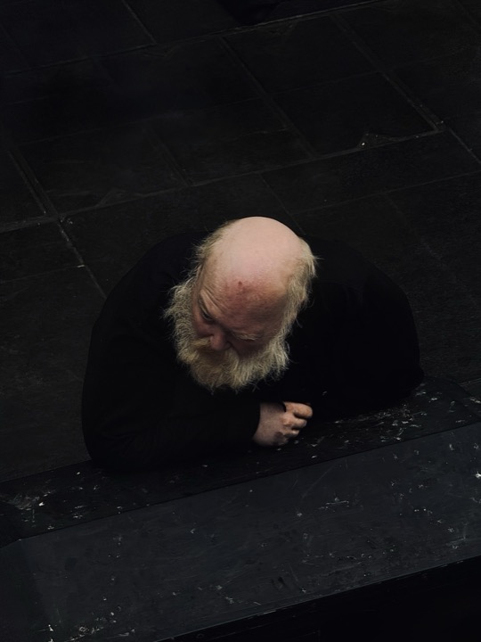 An overhead portrait of an older bearded man leaning on a dark surface.